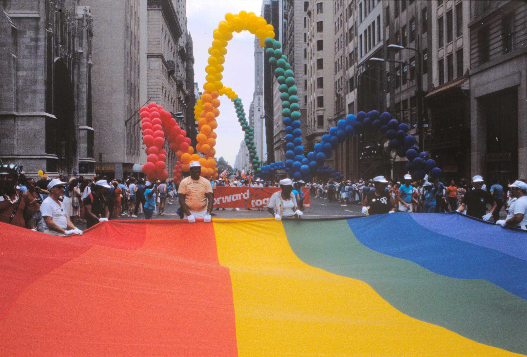 Click to view larger image Group of people holding a rainbow-striped banner across a city street. There is a parade behind them.
