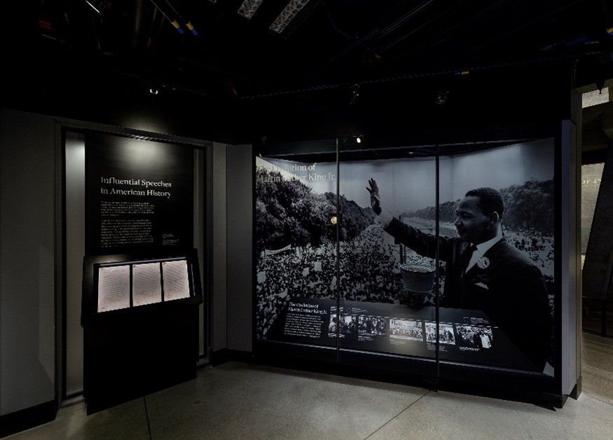 Photograph of wall panel with black and white photo in gallery next to case with three papers.