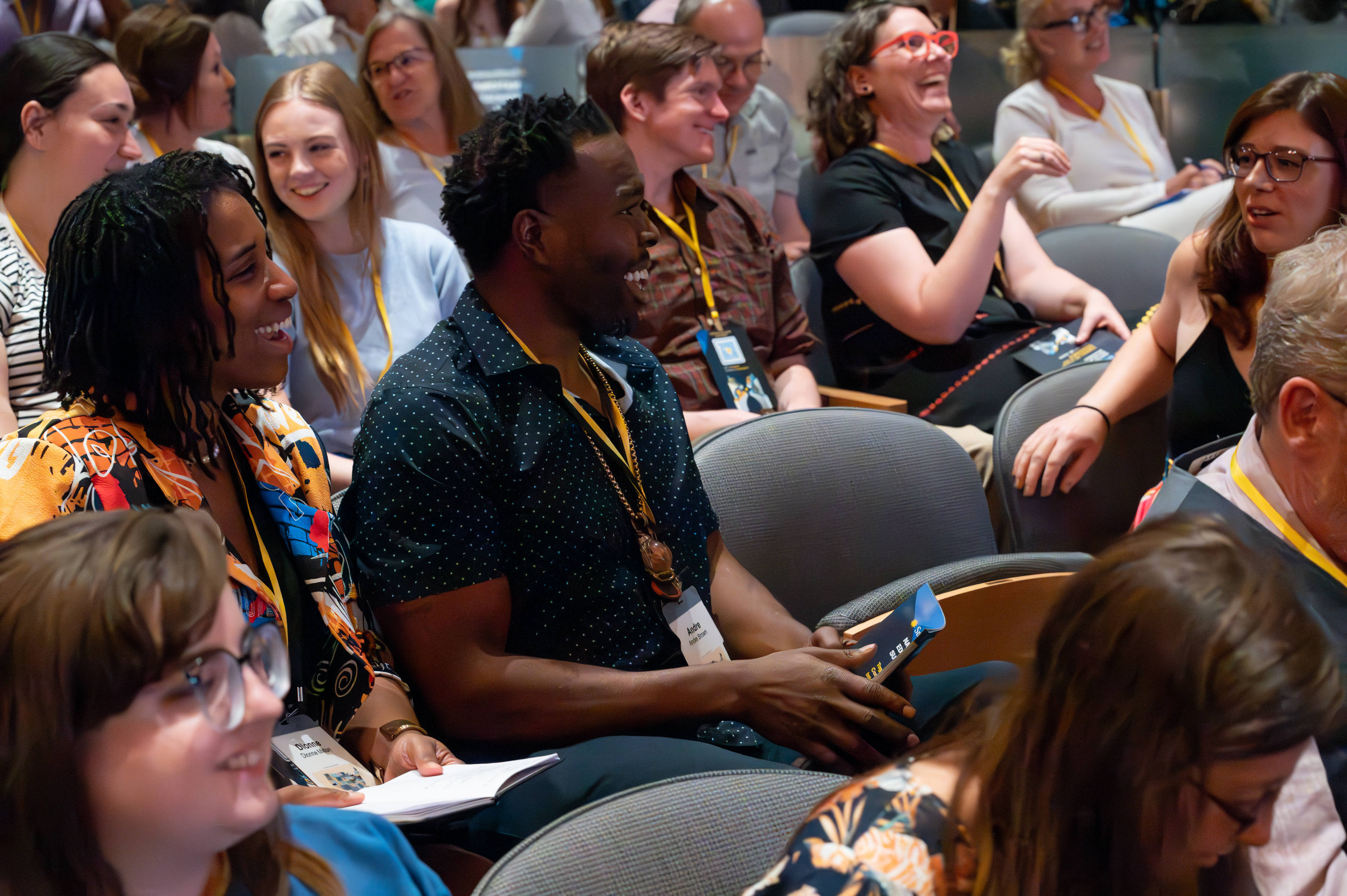 A group of people sitting in rows of chairs smile and converse with each other.