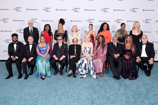 Two rows of 17 people dressed in black tie attire pose on a blue carpet