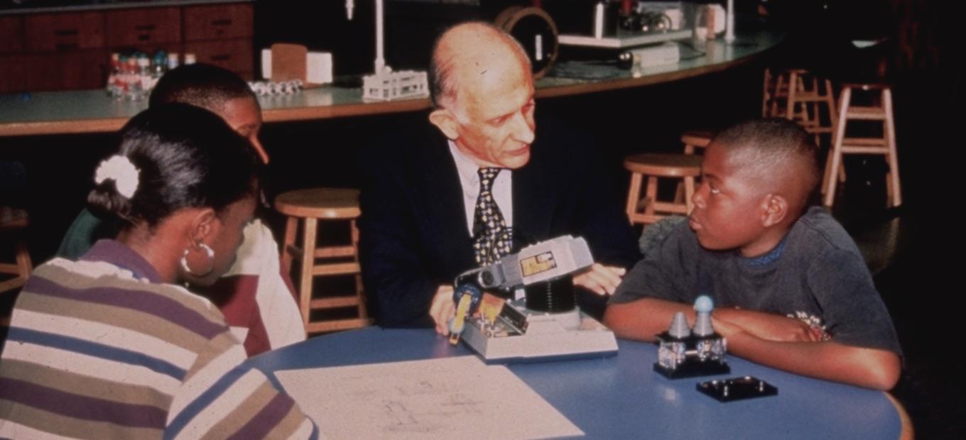 Older man in a suit sits at a round table with younger kids looking at microscope.