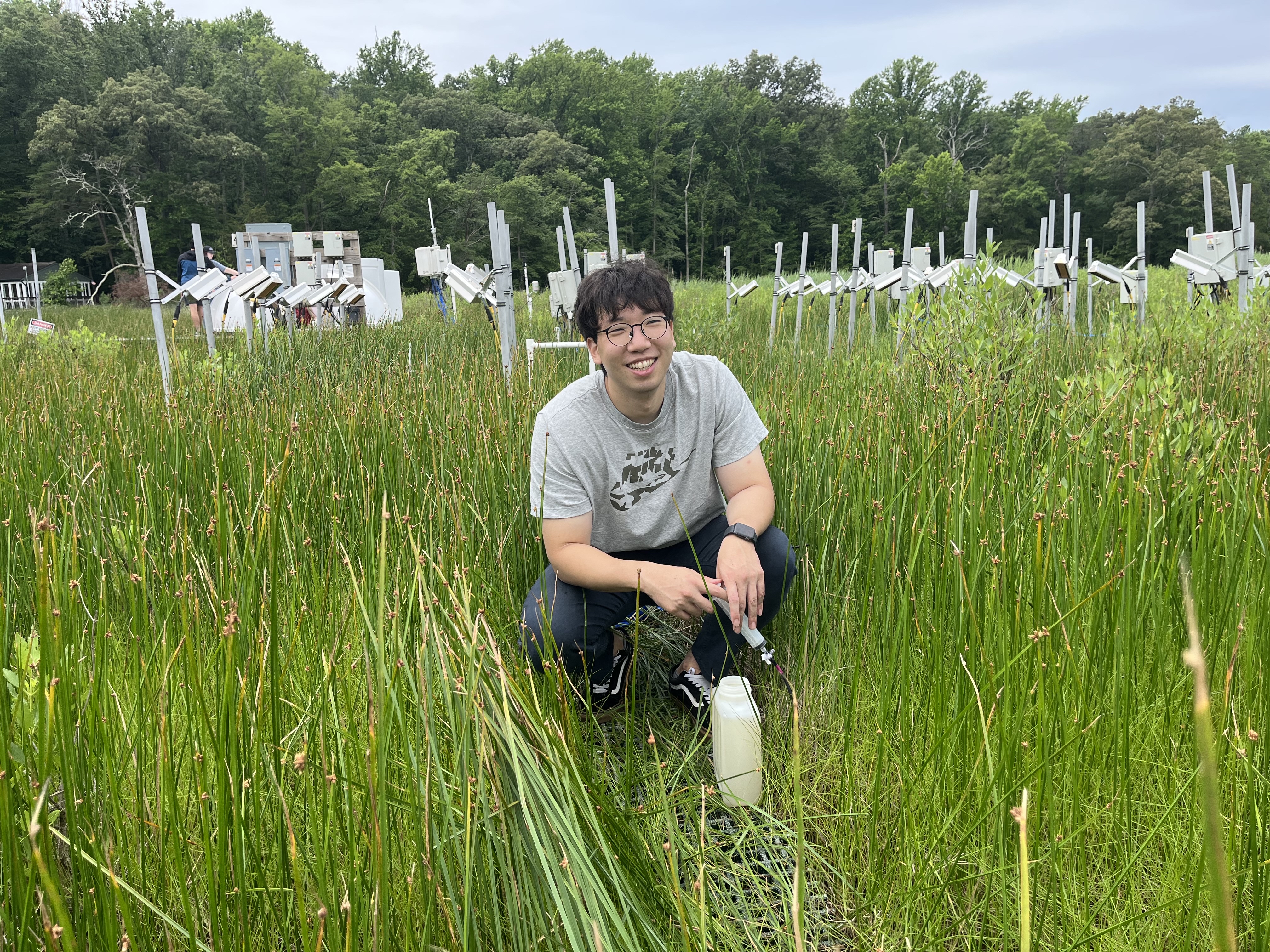 Man wearing glasses ans short sleeve shirt crouches down for a photo in a field of tall green grass.