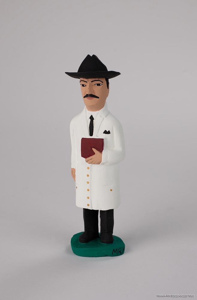 Small wooden figurine of a man in a long white lab coat and black hat holding a book.
