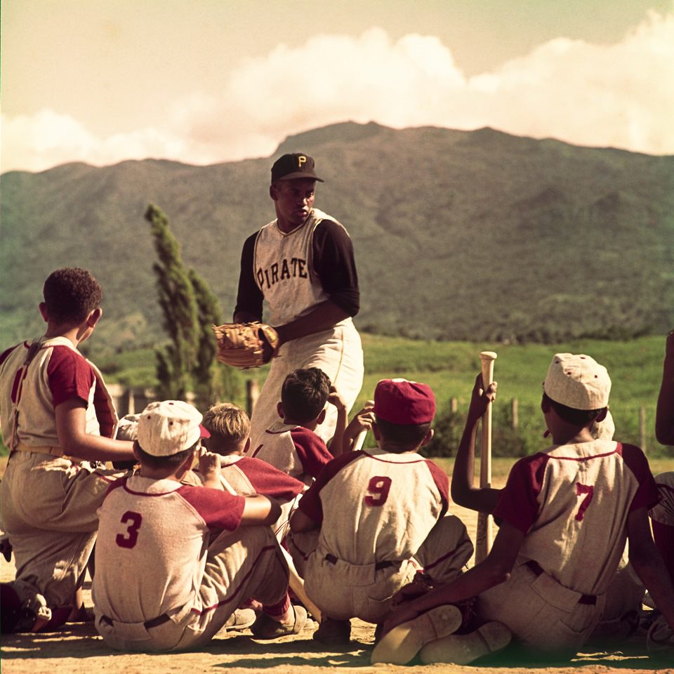 Man in baseball uniform stands above a group of young children whose backs are to the viewer