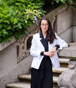 Woman smiling in black dress, white blazer, and glasses stands in front of stairwell.