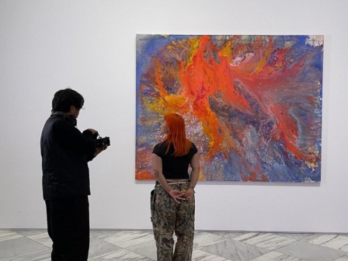 A woman stands looking at a piece of art on the wall while being filmed by another figure.