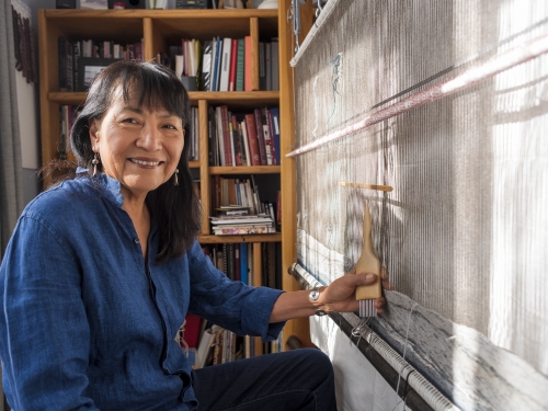 Woman with dark hair and a blue shirt, sitting in front of a loom.