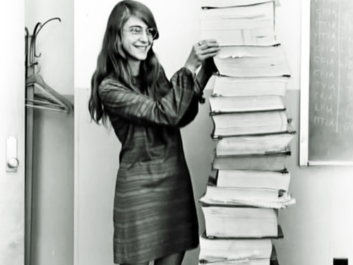 Black and white photo of a young woman standing next to stacked books and papers that are taller than her.