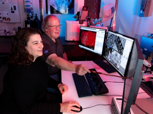 A man and a woman sit in front of three computer screens, pointing to digital images.