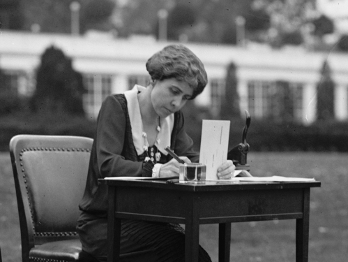 Black-and-white photograph of First Lady Grace Coolidge holding an absentee ballot envelope and pen while seated at a desk.