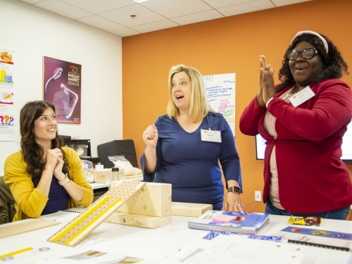 Three women stand around a table talking with education materials in front of them.
