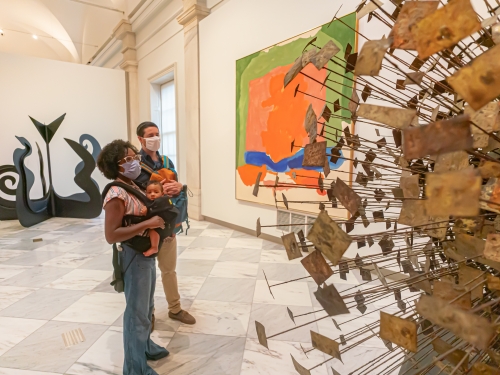 Two people carrying children look at sculpture in a museum