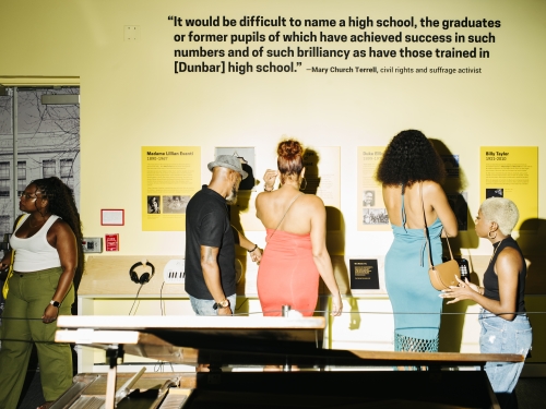 A group of four people tour a museum gallery with yellow walls, black labels on the wall and a piano.