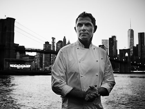 Black and white image of a man in a chefs coat standing in front of New York skyline.