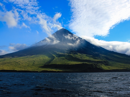 Volcano by the sea with wispy clouds floating around it