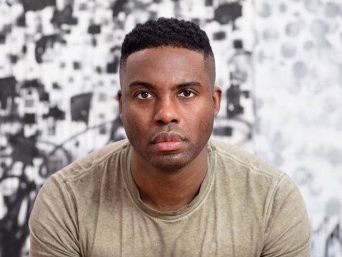 Man with dark skin and grayish t-shirt stands with hands clasped.