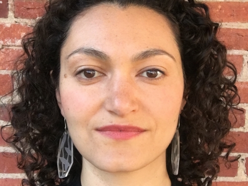 Picture of woman with dark curly hair from the shoulders up, standing against a brick background.