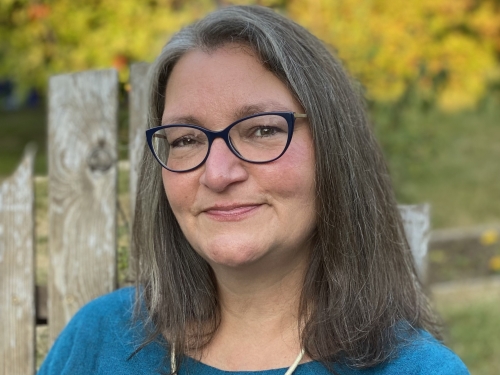 A woman with shoulder-length brown hair and gray highlights framing faces wears black glasses and sits in wooden chair outdoors.