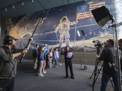 Group of children watch woman being filmed by cameramen in front of mural depicting space