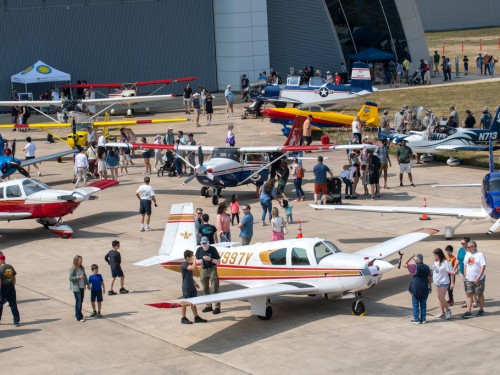 Overhead image of a crowd milling around small planes on a outdoors tarmac.