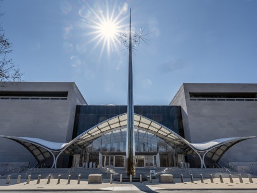 Exterior of light-colored blocky buildings with a designed glass entry and metal sculpture in front.