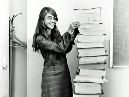 Black and white photo of a young woman standing next to a stack of books and papers that are taller than her.