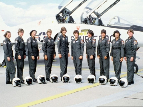 Image of a line of women standing in a row in matching gray jumpsuits with helmets sitting at their feet, in front of planes.