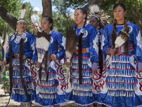 A group of four women wearing blue dresses with feathers stand in a line looking forward.