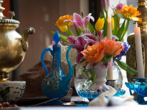 A table, viewed close up, that has been set with a vase of flowers, a blue-and-gold spouted jar, and candles