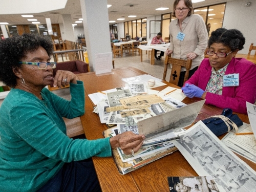 Two women sit opposite each other at wooden table, looking at archival books with black and white photos and news cutouts.