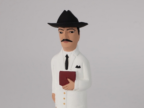 Small wooden figurine of a man in a long white lab coat and black hat holding a book.