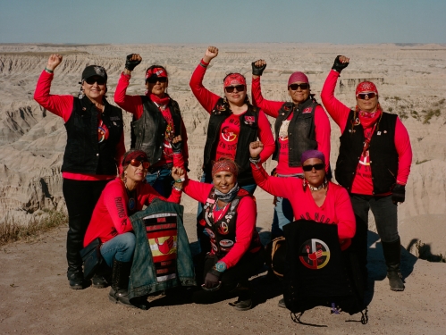 A group of women in red long-sleeved shirts raise one arm with fists, posing for a photo.
