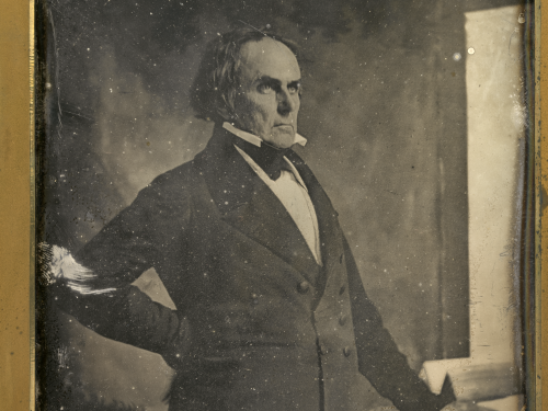 Aged black and white image of a man in a suit standing in front of a window.