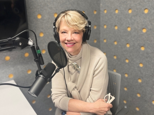 Woman with short blond hair and tan top sits in recording studio with headphones and a mic in front of her.