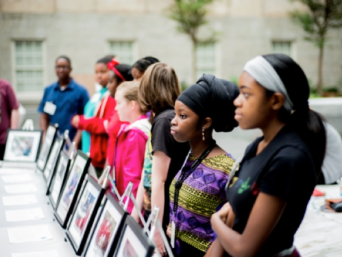 Image of a row of teenagers standing in front of propped up photographs.
