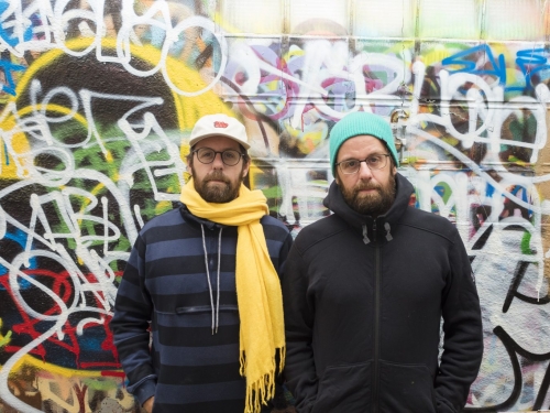 Two men in dark puffy jackets and glasses stand against wall of colorful grafitti.