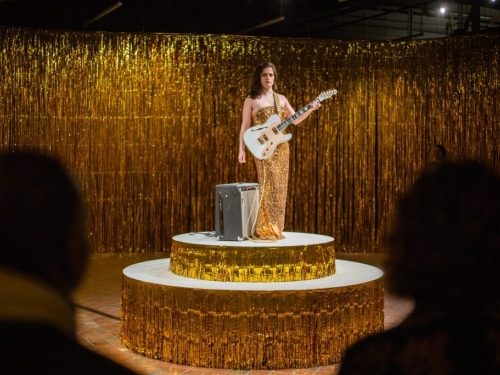 Woman in gold dress holding guitar on dais