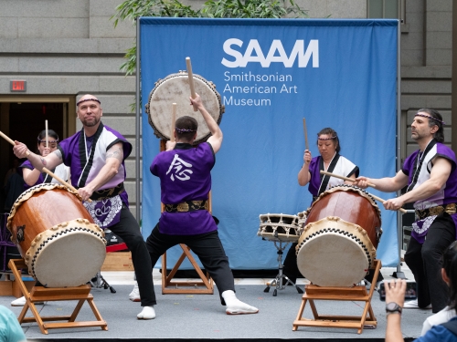 Three men and a woman stand on a stage playing large drums in a museum atrium.