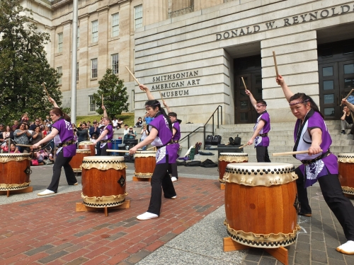 several people play large drums enthusiastically in from of a building