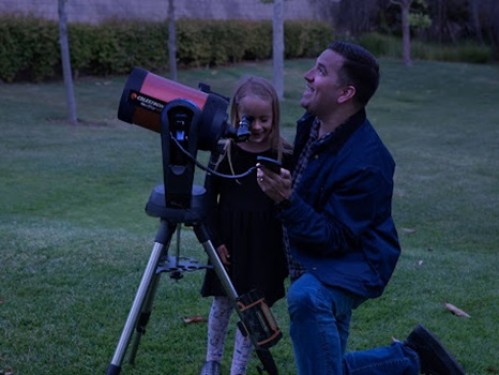 An older man kneels next to a telescope and young girl looking at the sun.