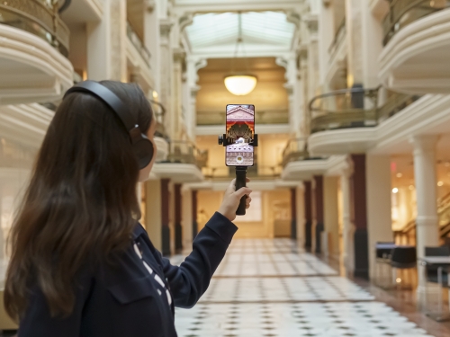 Girl with brown hair and black headset holds up a phone with an augmented reality graphic on it.