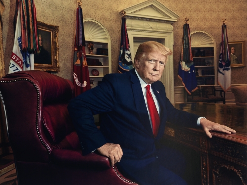 Man in a black suit and red tie sits a dark wooden table in the Oval Office.