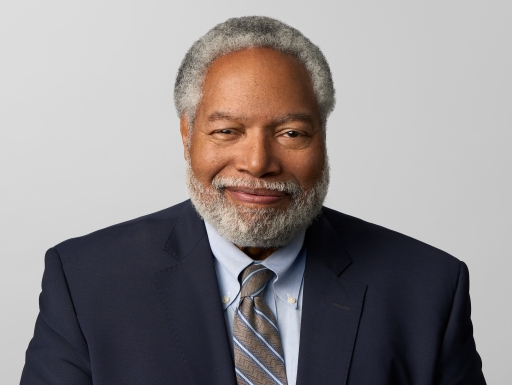 Man with short silver and black hair, wearing a dark suit and light blue shirt, smiles toward camera/