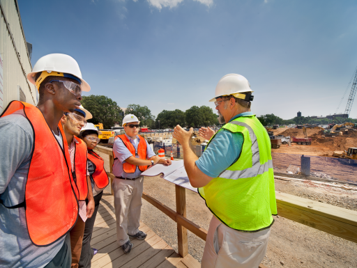 group of people at a construction site looking at a man speaking everyone is wearing reflective vests and hard hats