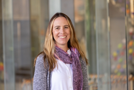 Woman in a white top, gray cardigan and purple scarf smiles outside a building with glass siding.