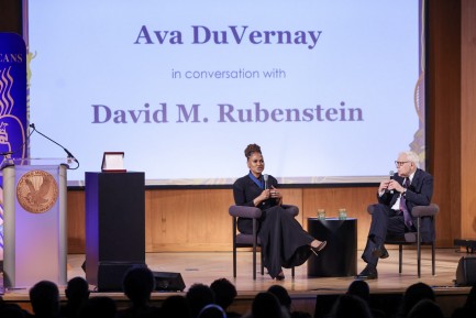 A woman and a man sit in two chairs on a stage speaking into mics.