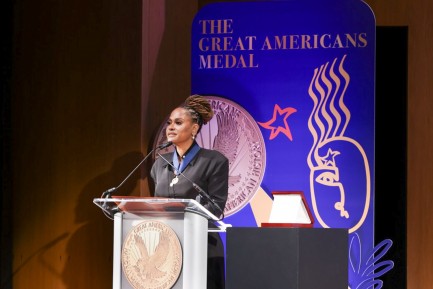 A woman wearing a black evening gown stands in front of a podium delivering remarks.