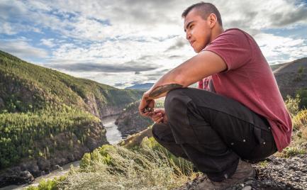 still image of squatting man overlooking a river