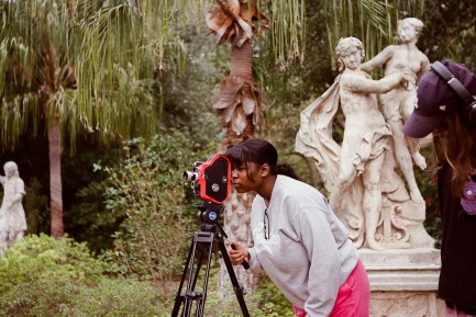 A girl in a light grey sweatshirt looks into large camera on tripod in outdoor sculpture garden.