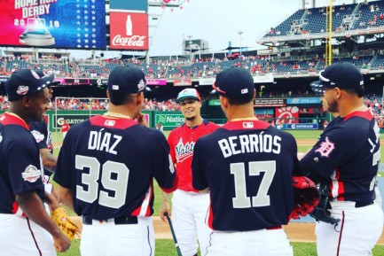 Four baseball players with their backs to the viewer talk to another player facing the camera in a baseball stadium